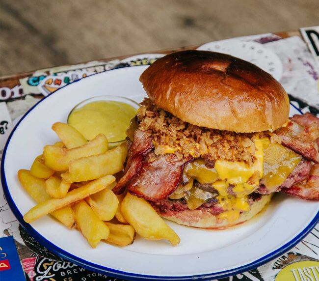 Apetecible Burger Doble Cheddar de la hamburguesería Garua Bar, con carne de chuleta de 360gr, cocinada a la plancha, acompañada por una generosa capa de doble queso cheddar fundido, doble de bacon crujiente, cebolla frita dorada y unas patatas de guarnición en la hamburguesería Garua Bar de Donostia-San Sebastián