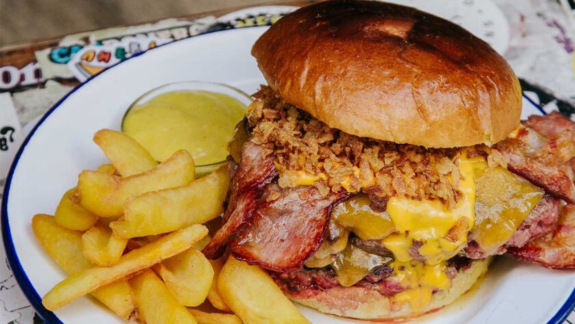 Apetecible Burger Doble Cheddar de la hamburguesería Garua Bar, con carne de chuleta de 360gr, cocinada a la plancha, acompañada por una generosa capa de doble queso cheddar fundido, doble de bacon crujiente, cebolla frita dorada y unas patatas de guarnición en la hamburguesería Garua Bar de Donostia-San Sebastián