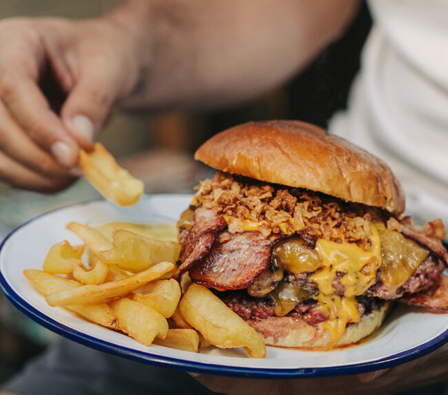 Apetecible Burger Doble Cheddar de la hamburguesería Garua Bar, con carne de chuleta de 360gr, cocinada a la plancha, acompañada por una generosa capa de doble queso cheddar fundido, doble de bacon crujiente, cebolla frita dorada y unas patatas de guarnición en la hamburguesería Garua Bar de Donostia-San Sebastián