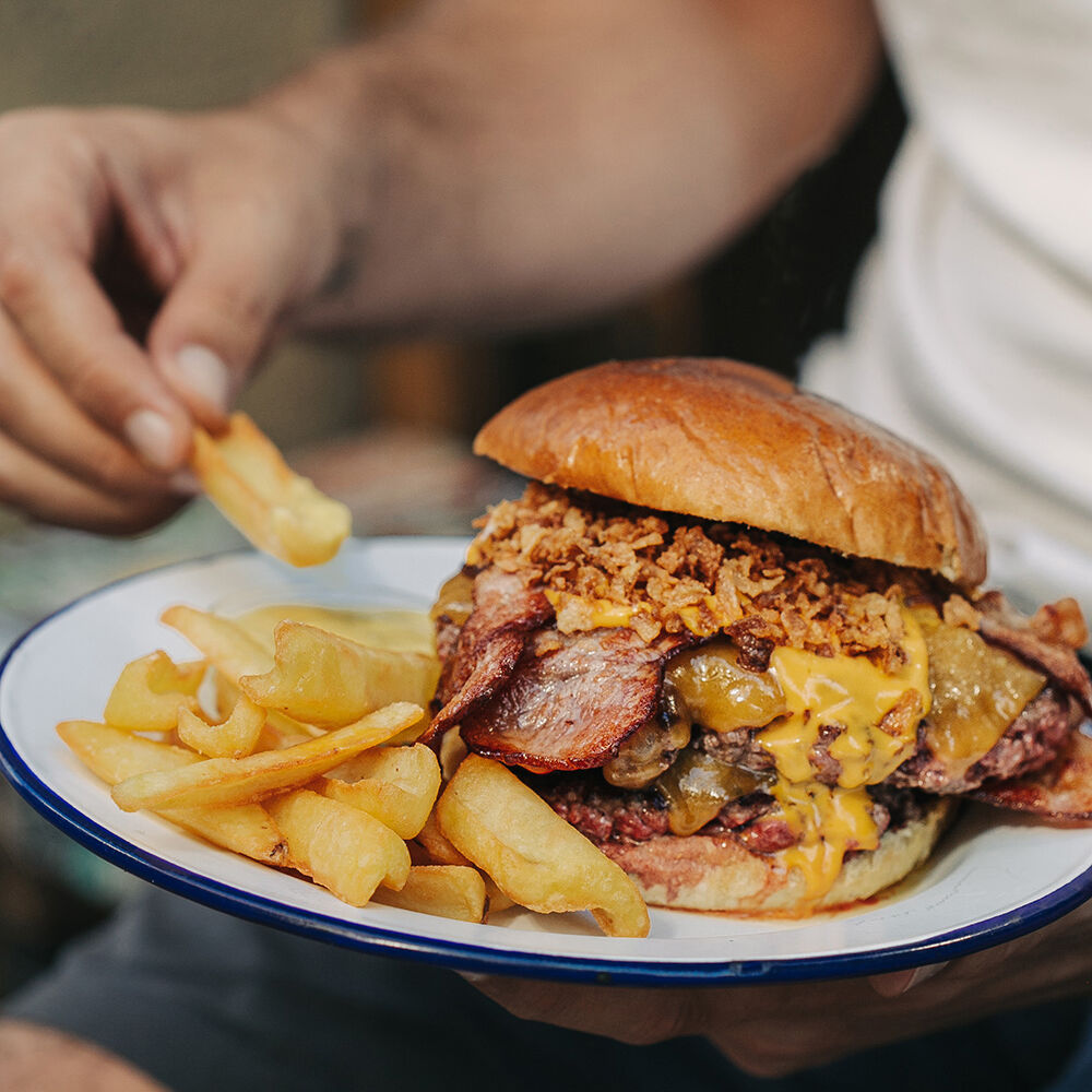 Apetecible Burger Doble Cheddar de la hamburguesería Garua Bar, con carne de chuleta de 360gr, cocinada a la plancha, acompañada por una generosa capa de doble queso cheddar fundido, doble de bacon crujiente, cebolla frita dorada y unas patatas de guarnición en la hamburguesería Garua Bar de Donostia-San Sebastián